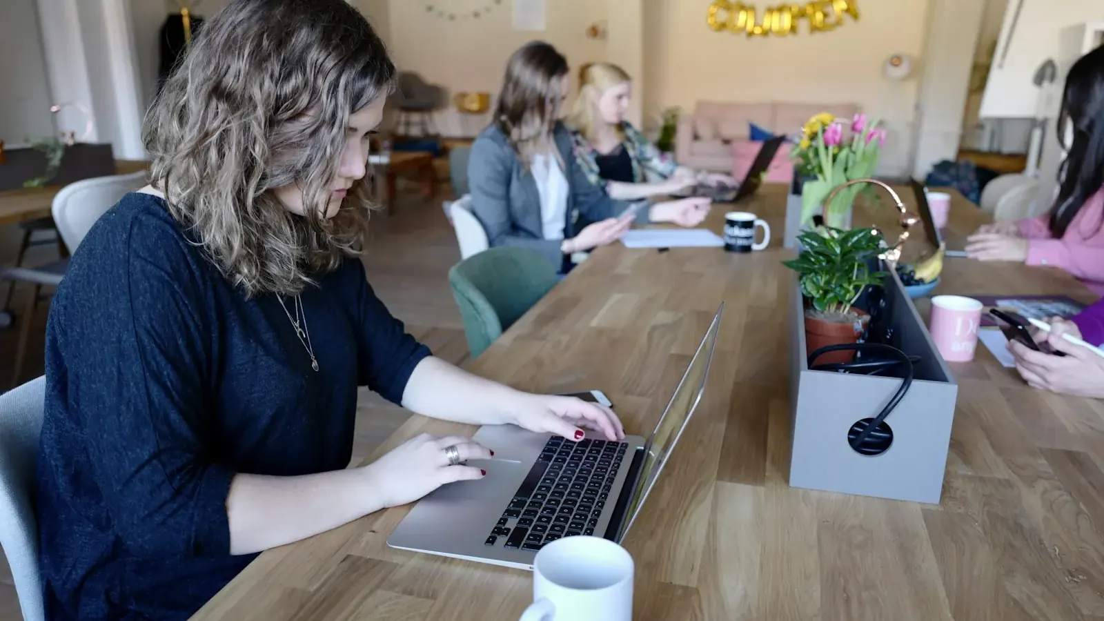 Eine junge Frau tippt konzentriert am Laptop an einem Gemeinschaftstisch im Coworking-Space.