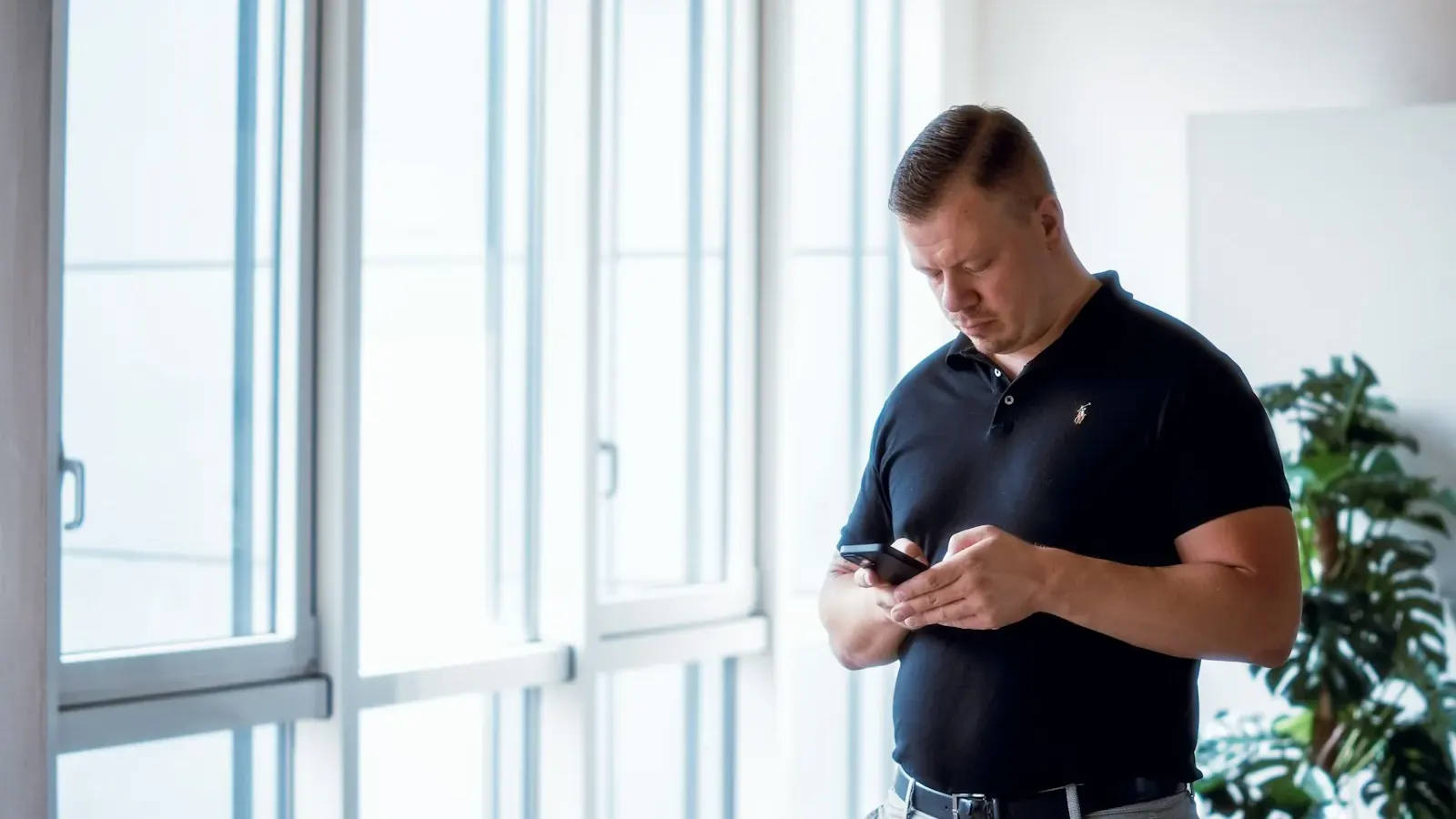 Ein Mann im Poloshirt schreibt in einem hellen Büro Nachrichten auf dem Smartphone.