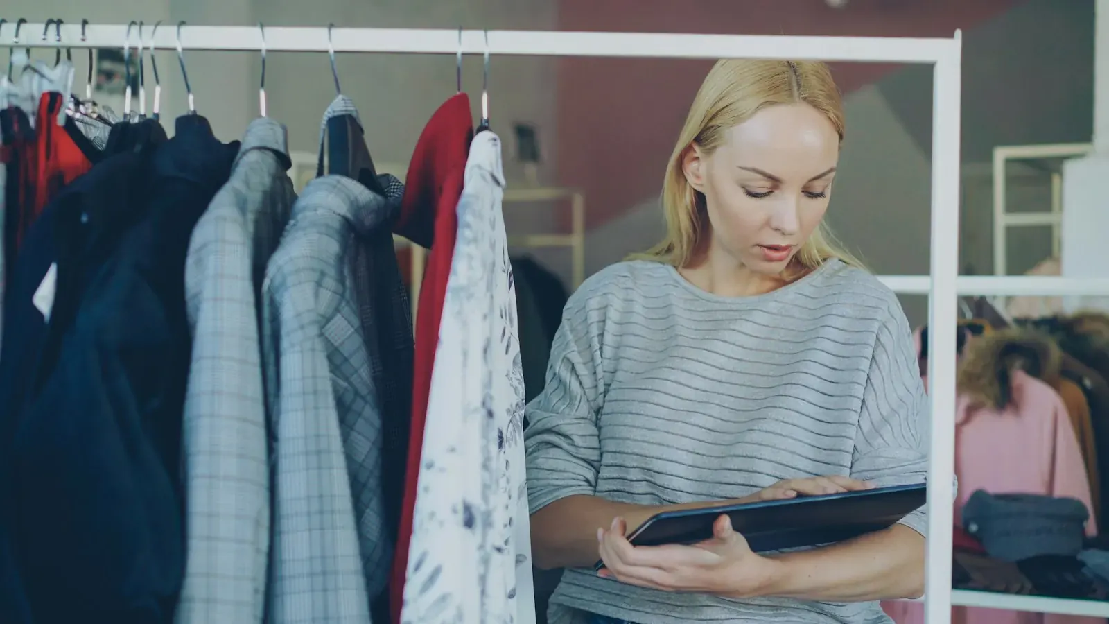 Eine Boutique-Inhaberin steht mit einem Tablet zwischen Kleiderstangen und plant ihren Onlineshop.