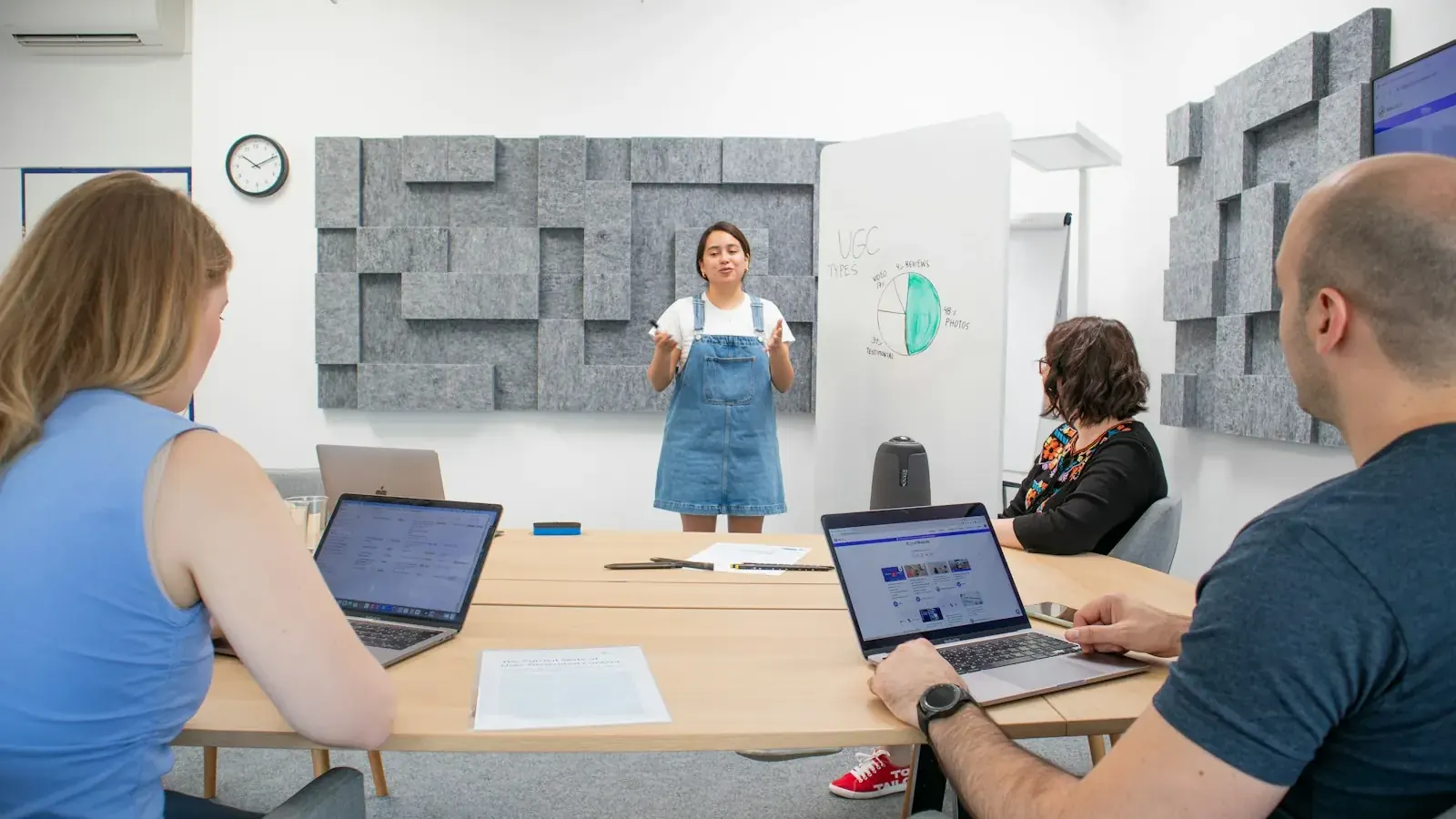 Eine junge Frau präsentiert ihrem Team an einer Tafel, auf dem Bildschirm ein Website-Entwurf.