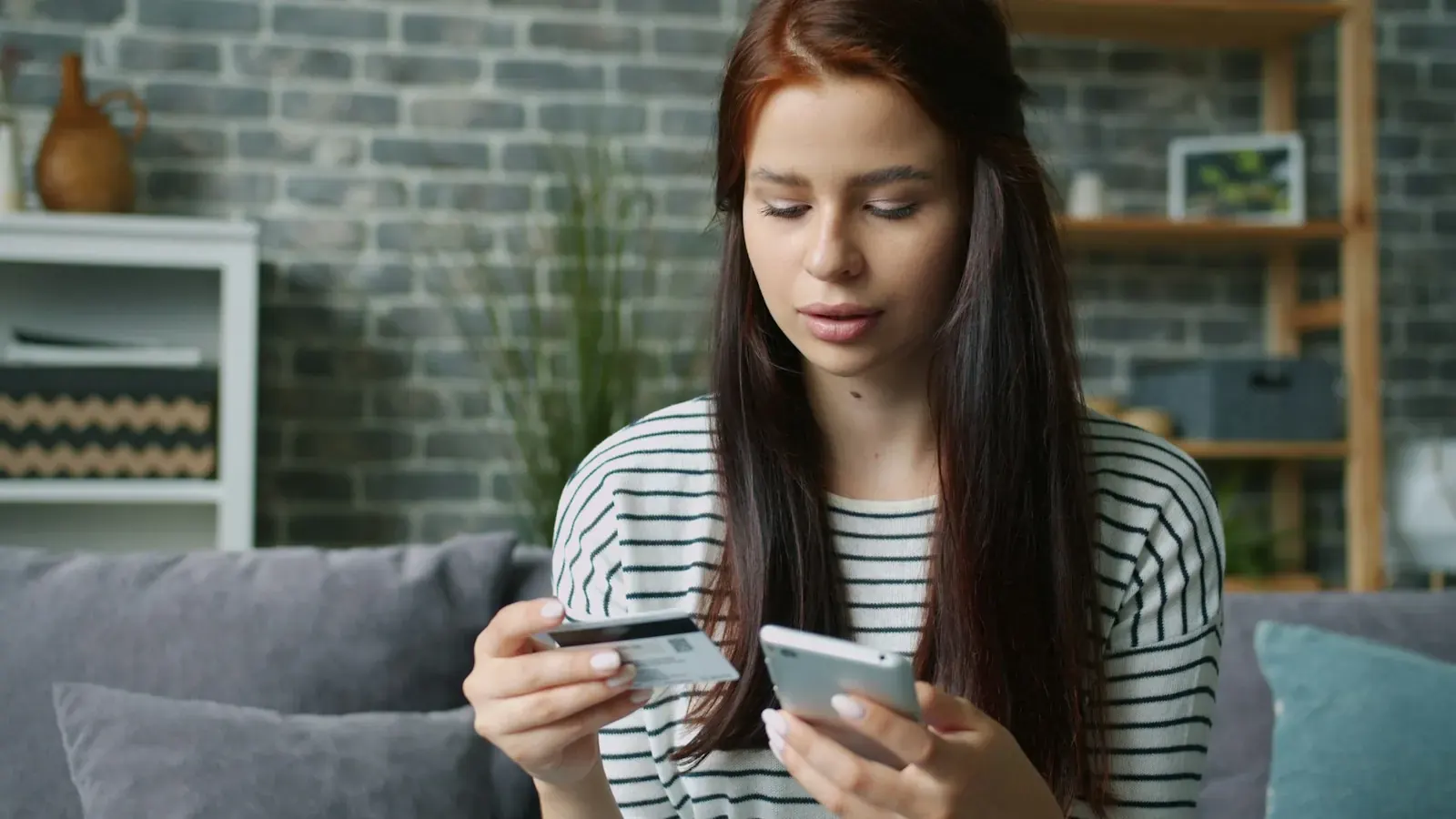 Eine junge Frau vor einer Backsteinwand bezahlt mit Bankkarte und Smartphone online.