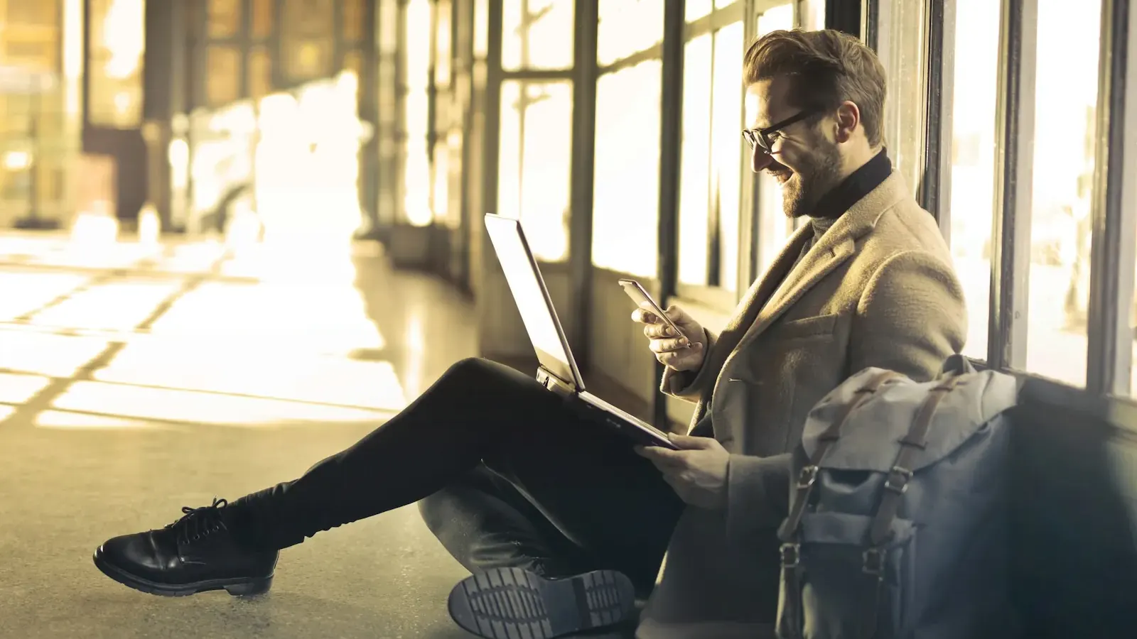 A founder sits on the floor of a sunny lobby with a laptop and phone.