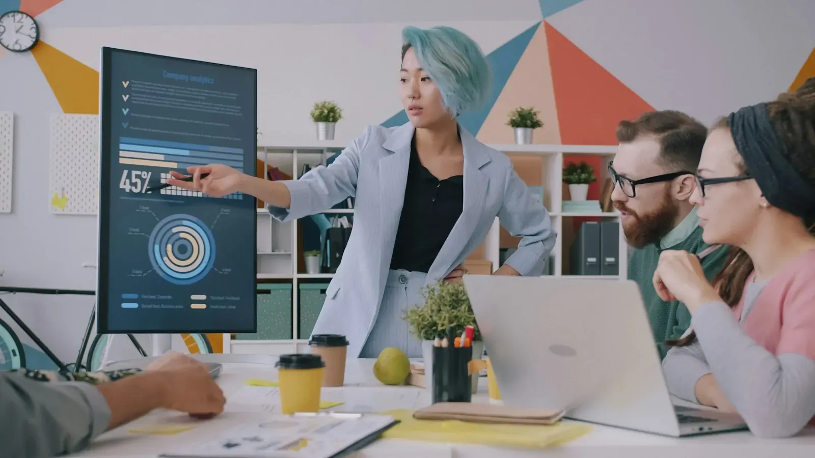 A woman shows her team a dashboard with charts on a large screen.