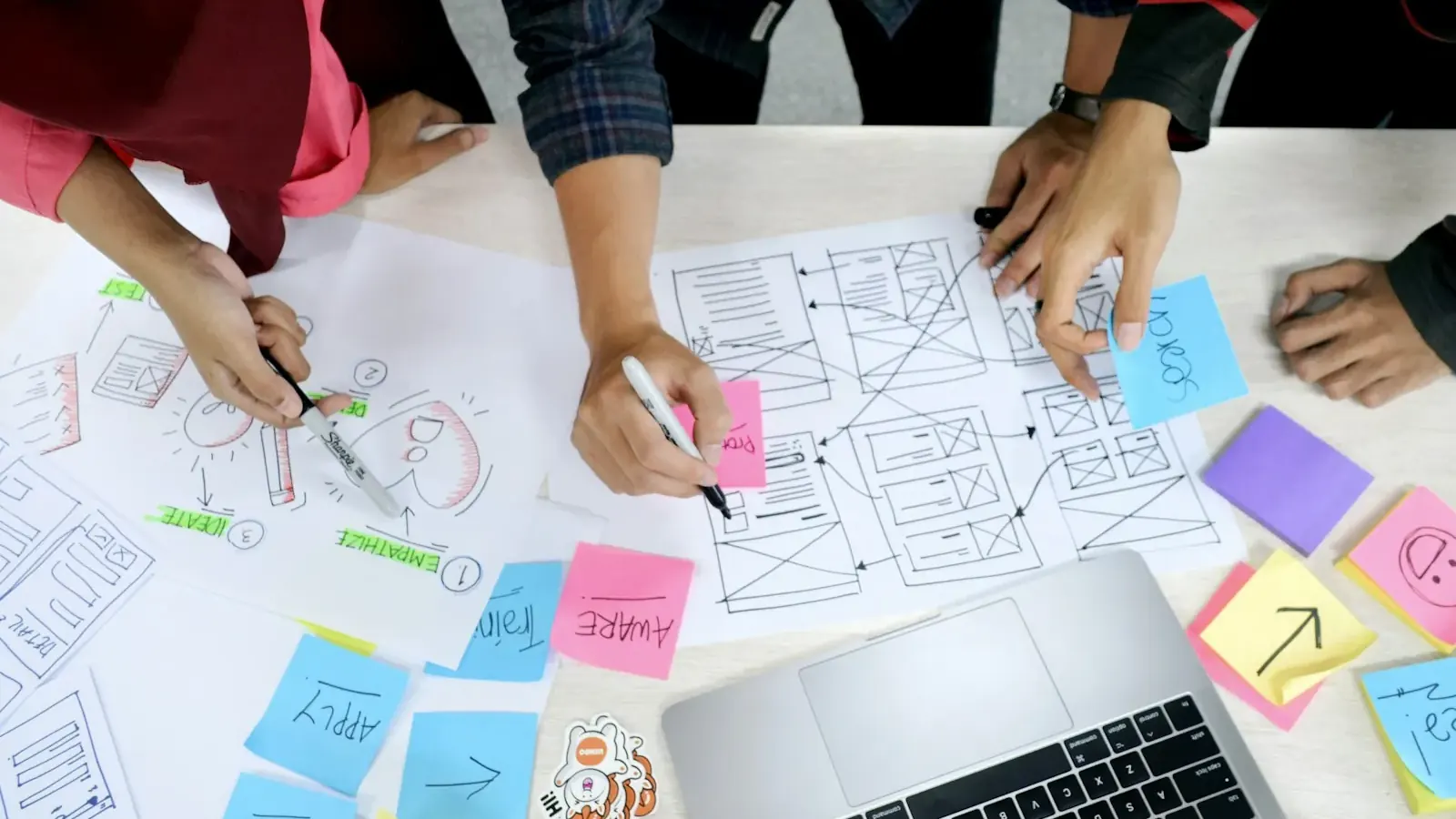 From above, a team sketches app screen wireframes with sticky notes beside a laptop.