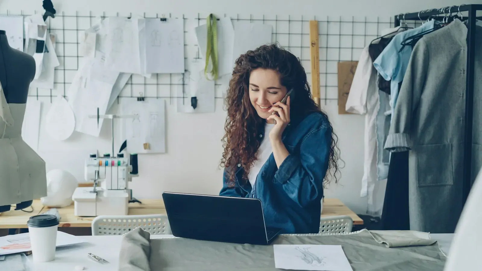A clothing designer talks on the phone at her laptop while planning an online store.