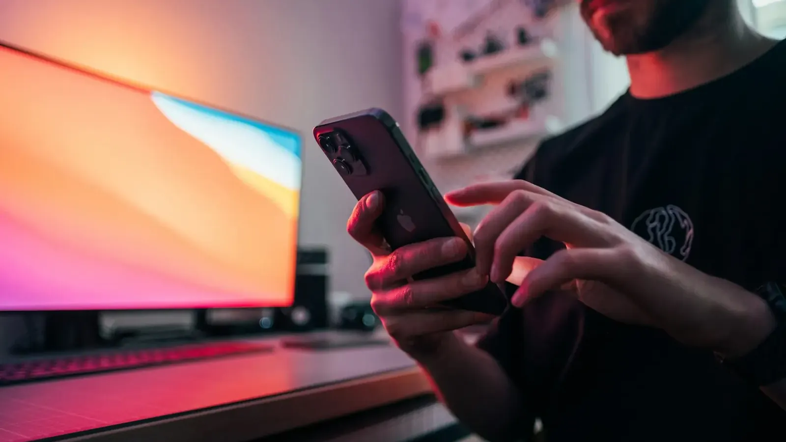 A man uses a smartphone in front of a bright pink and blue monitor.