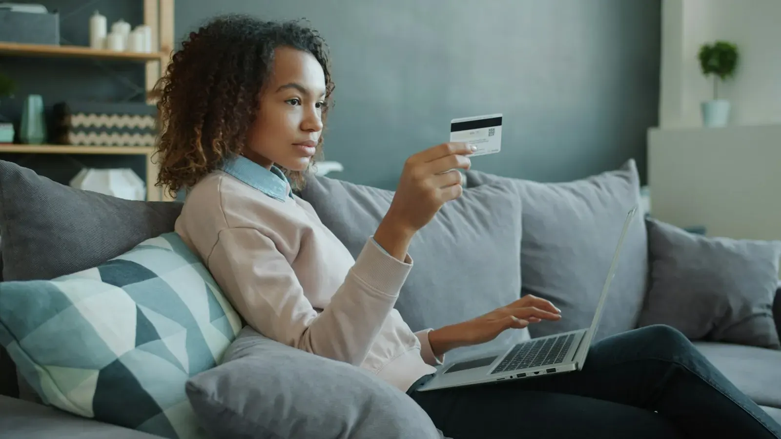 A young woman on a sofa pays by card online through a laptop.