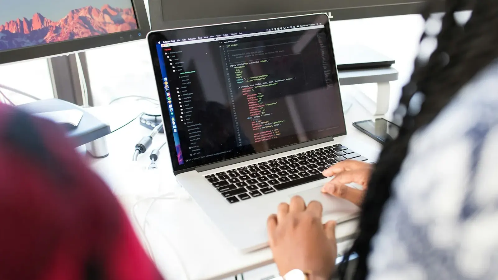 A developer's hands type code on a laptop beside a colleague in a bright office.