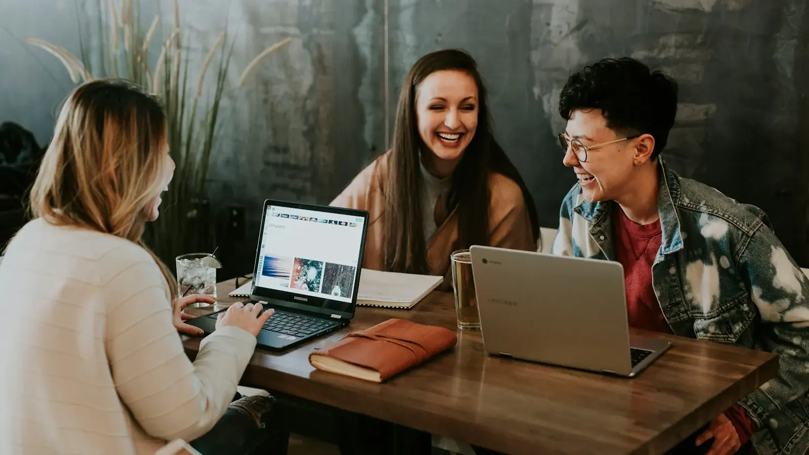 Three colleagues laugh at their laptops in a cafe, with a website on screen.