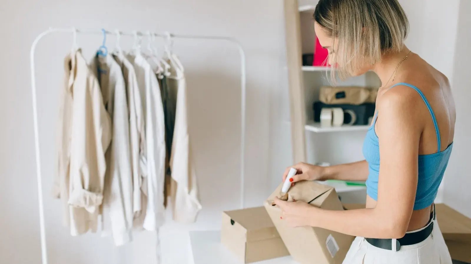 Una dueña de tienda de ropa empaqueta un pedido en una caja junto a un perchero.