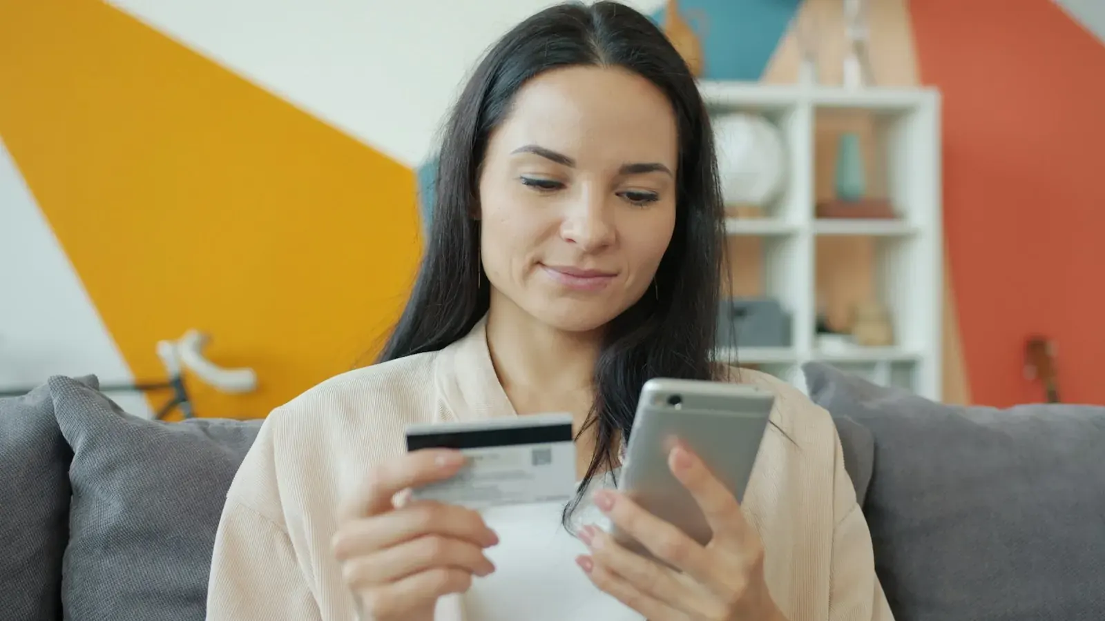 Una chica morena con tarjeta bancaria y móvil paga online ante un portátil.