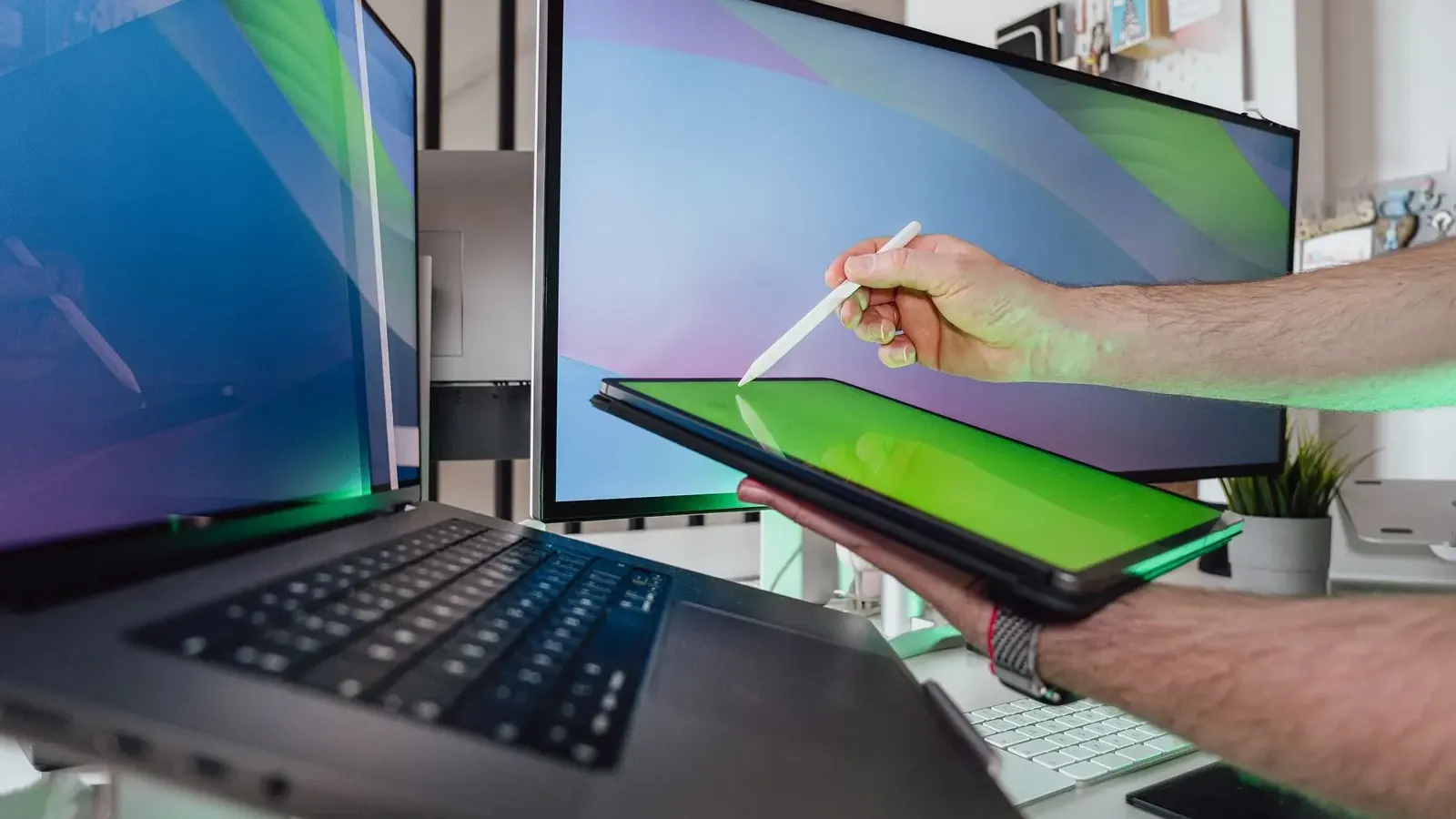 Dłoń z rysikiem nad tabletem przy laptopie i monitorach w zielonym podświetleniu.