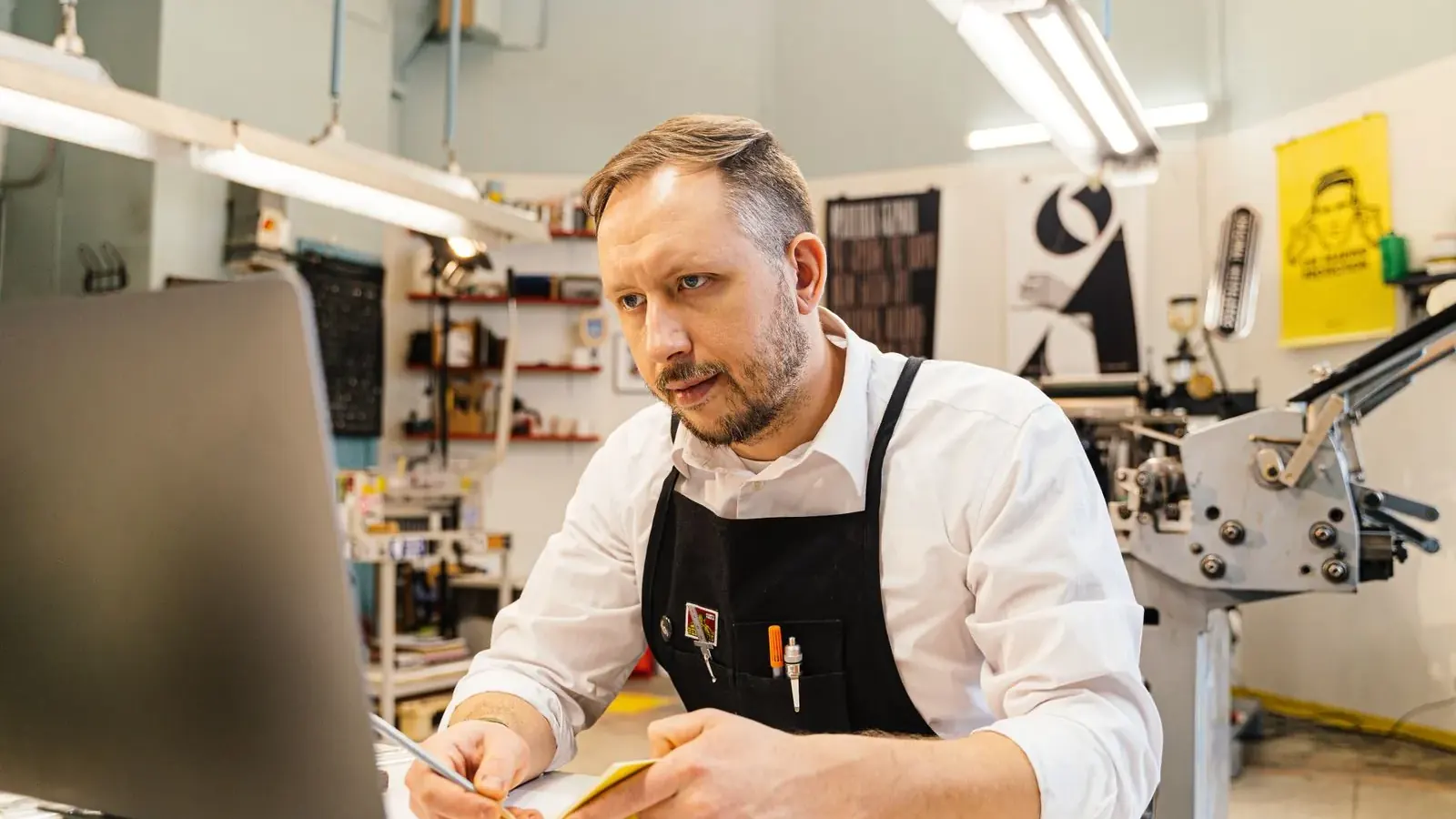 Rzemieślnik w fartuchu pracuje przy laptopie w swojej pracowni i patrzy w obiektyw.