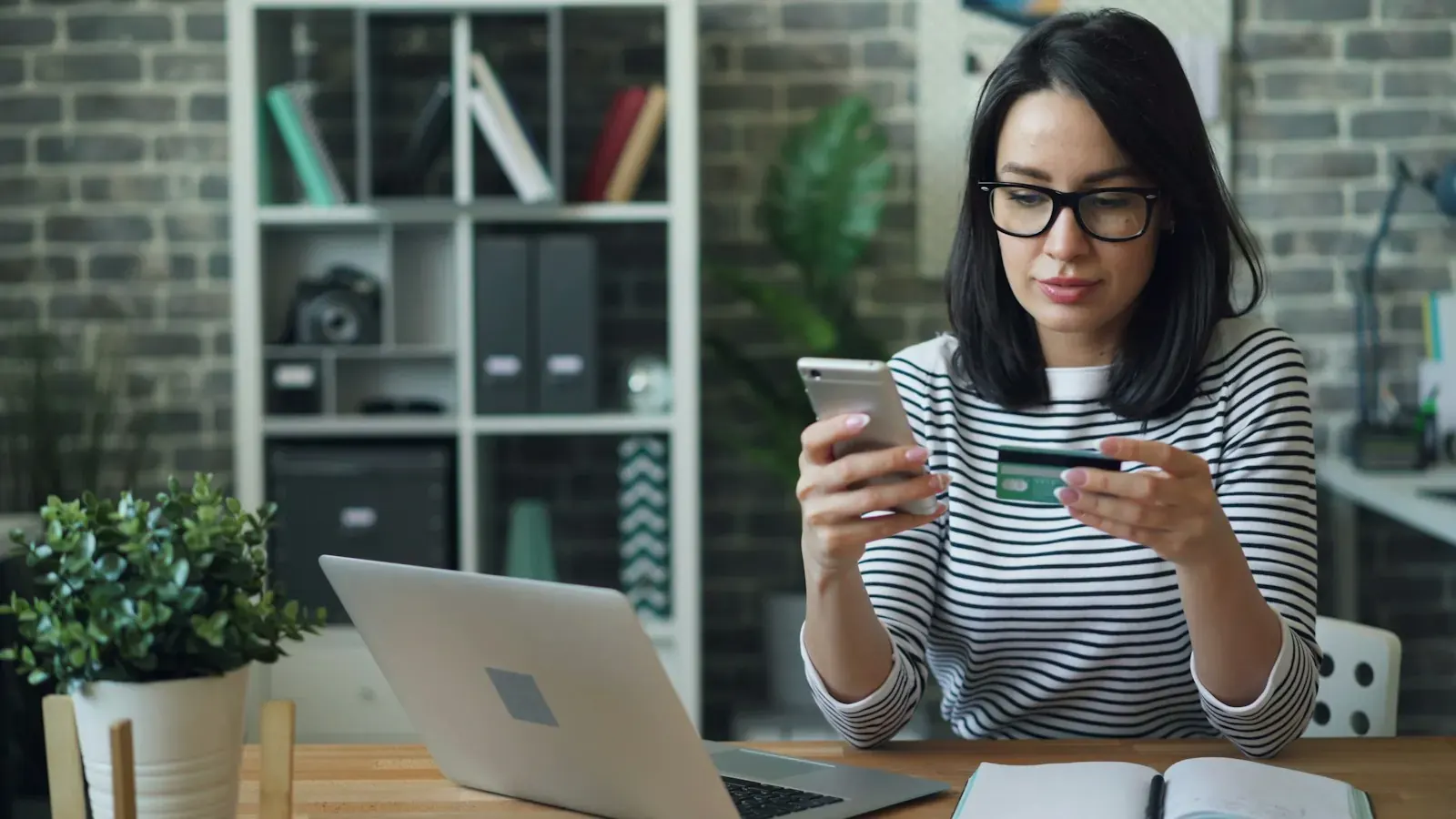 Młoda kobieta z kartą bankową i smartfonem płaci online przy laptopie.