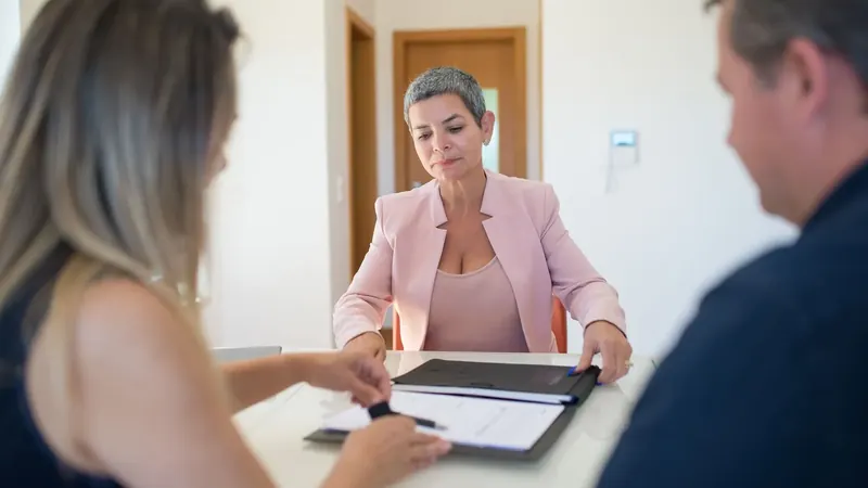 Uma consultora ajuda uma cliente a assinar documentos à mesa de um escritório.