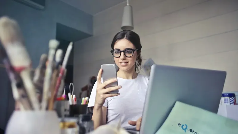 Uma jovem de óculos usa um telemóvel junto a um portátil num estúdio de design.