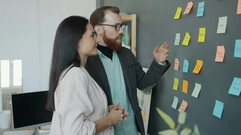 Um homem e uma mulher discutem o plano do projeto junto a uma parede de notas.