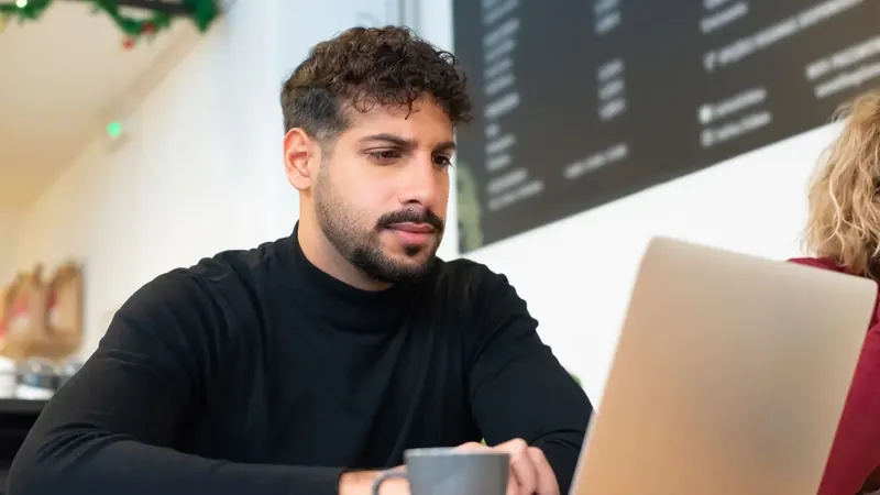 Um homem lê com atenção num portátil num café, com uma chávena de café ao lado.
