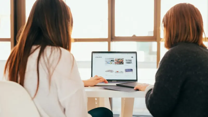 Duas jovens junto a uma janela solarenga analisam um site num portátil.